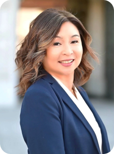 Smiling woman with wavy shoulder-length brown hair wearing a navy blazer over a white top, standing outdoors with a blurred background.