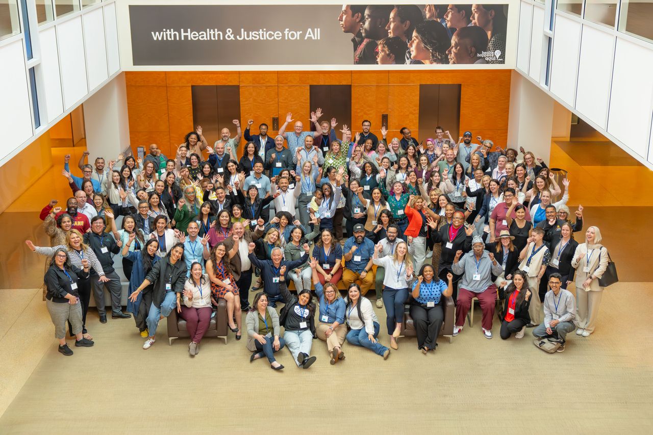 Large group of CACHI network members gathered in a building atrium, cheering and raising their hands, beneath a banner reading 'with Health & Justice for All'.