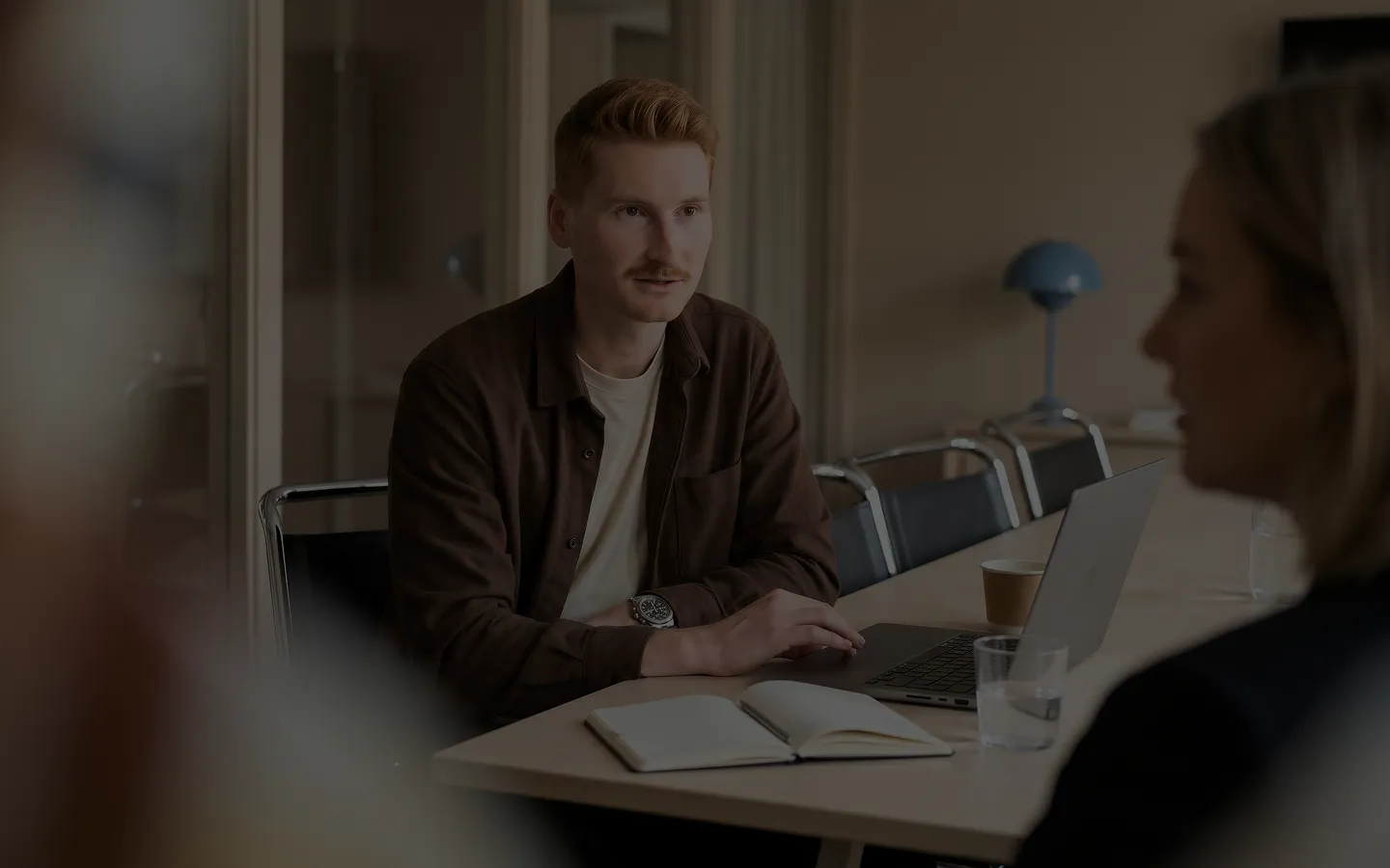 A man with short red hair and a mustache wearing a brown jacket sits at a table with an open notebook and a laptop, engaged in conversation with two blurred individuals in a meeting room.