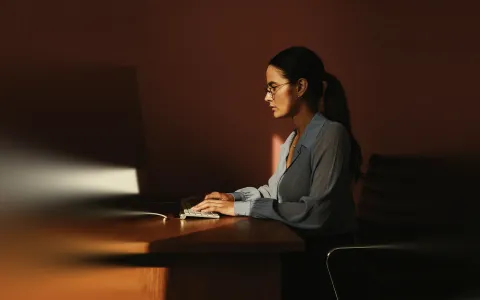 Woman with glasses and long hair tied back sitting at a wooden desk typing on a small keyboard, illuminated by natural light against a dark background.