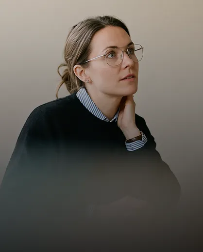 Woman with light brown hair tied back wearing large round glasses, a striped shirt under a black sweater, resting her neck with one hand and looking upward thoughtfully.