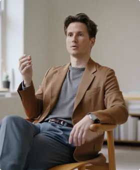 A man with light skin and dark hair wearing a brown blazer and gray shirt sits on a wooden chair in a modern indoor setting, gesturing with one hand while speaking.