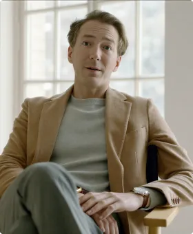 A man with light skin and wavy brown hair is sitting on a wooden chair near large windows, wearing a brown blazer over a light gray shirt and gray pants, with one leg crossed and hands resting on his lap.