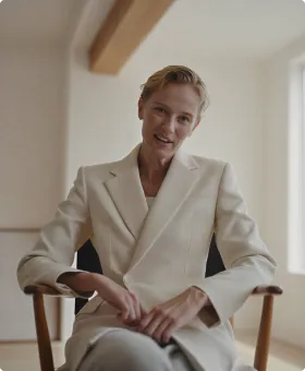 A person with short blond hair wearing a light beige suit jacket, sitting on a wooden chair with a relaxed posture in a softly lit room with neutral walls and a window.