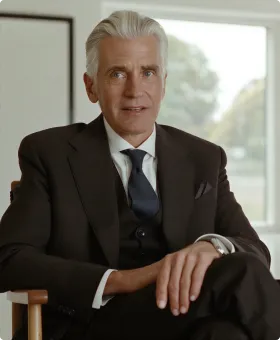 An older man with white hair wearing a black suit, white shirt, and dark tie, sitting on a wooden chair with a window showing a blurred outdoor view behind him.