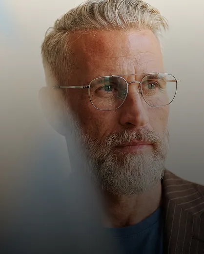 Close-up of an older man with light skin, gray hair, and a beard wearing metal-framed glasses and a brown striped blazer, looking thoughtfully to the side.
