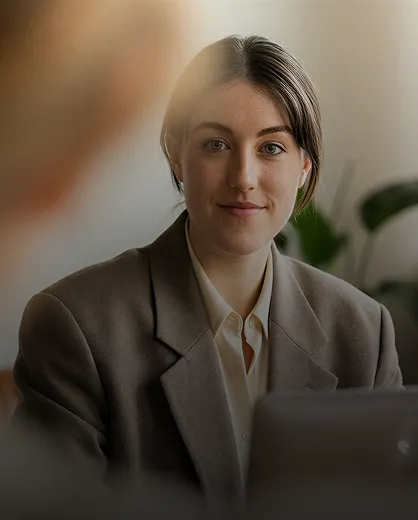 A woman with light skin and brown hair parted in the middle, wearing a brown blazer and white shirt, looking forward with a slight smile in a softly lit indoor setting.