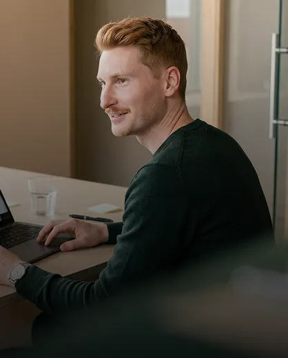 Young man with short reddish hair and a slight mustache sitting at a desk using a laptop, smiling and looking to his left.