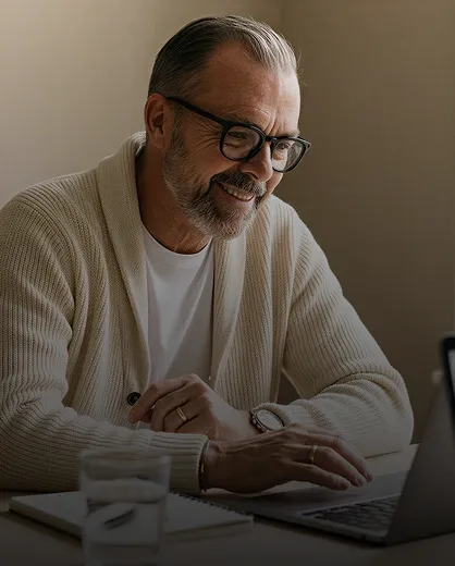 Smiling middle-aged man with glasses and gray beard wearing a white sweater, working on a laptop at a desk with a notebook and glass of water.