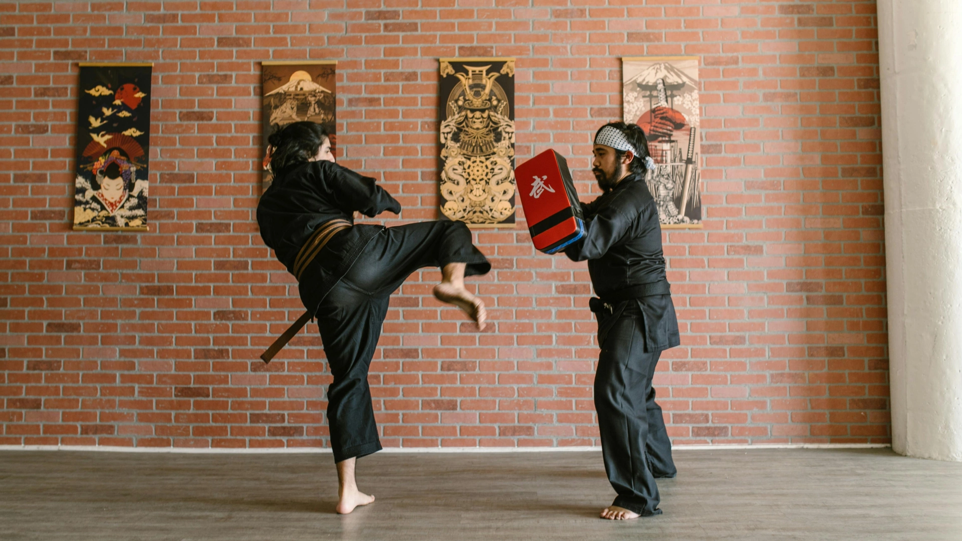 Martial arts student practicing a kick on a pad held by an instructor in a traditional dojo