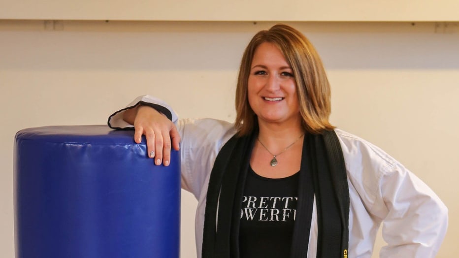 Smiling instructor in a white uniform and black belt leaning on a blue heavy bag
