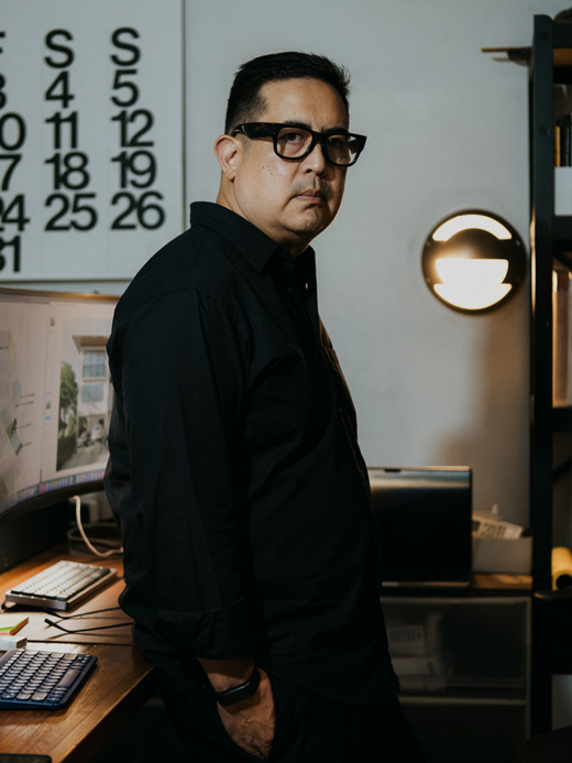 A man wearing black framed glasses and a black shirt stands with one hand in his pocket in a dimly lit office space, with computer monitors, a desk, and a round wall light in the background.