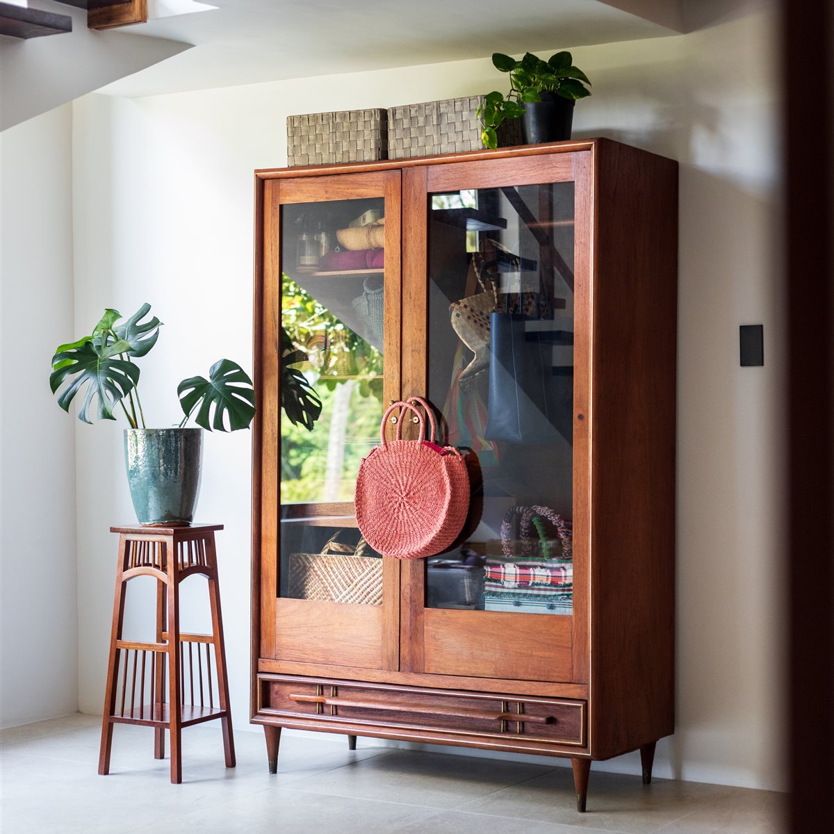 Wooden cabinet with glass doors displaying woven baskets, bags, and folded fabrics, with a red round woven bag hanging on the door and a potted plant on top, next to a tall wooden plant stand holding a potted monstera plant in a greenish-blue textured pot, all set on a tiled floor against a light-colored wall.