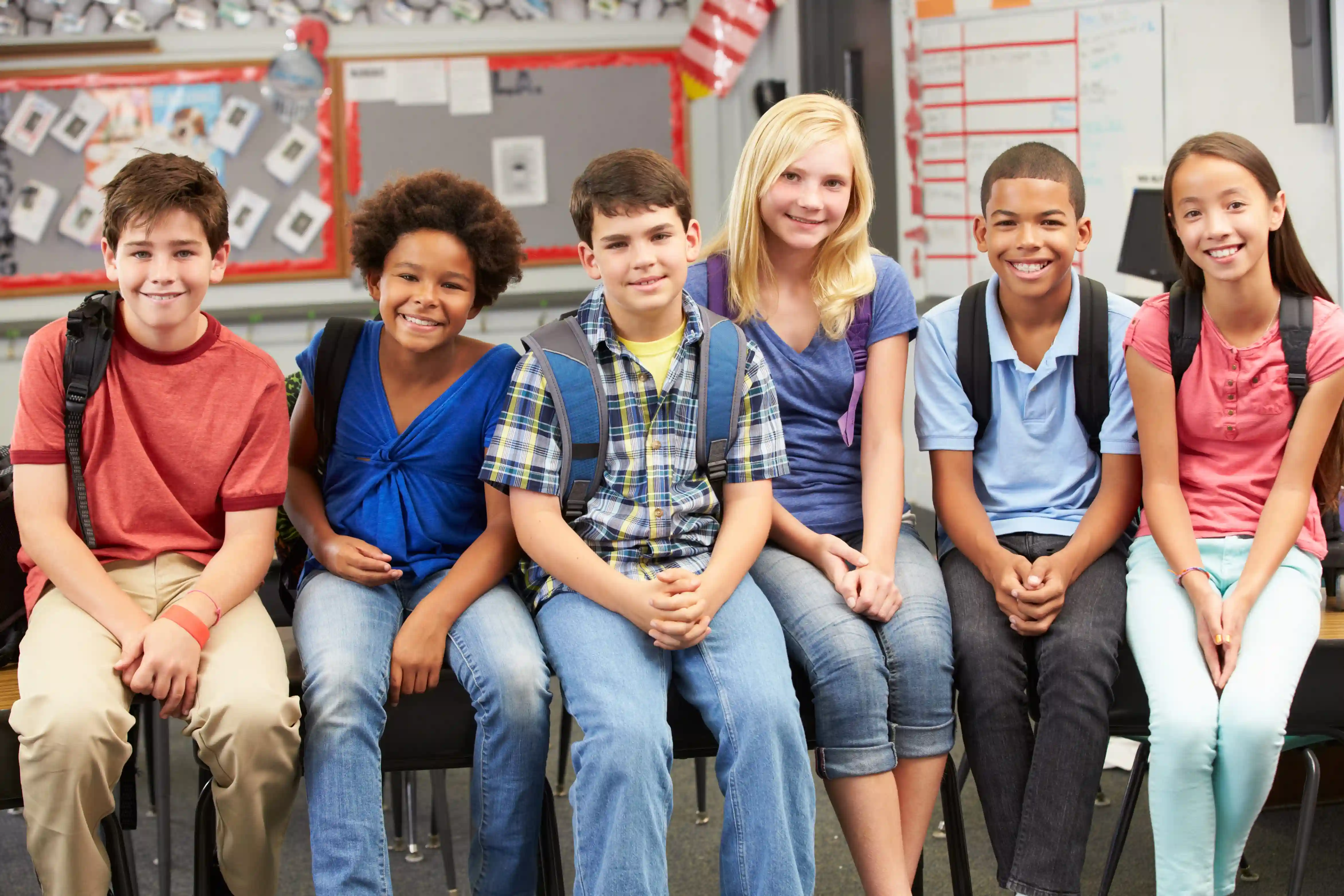 Four diverse middle school students sitting side by side in a classroom, wearing backpacks and smiling at the camera.