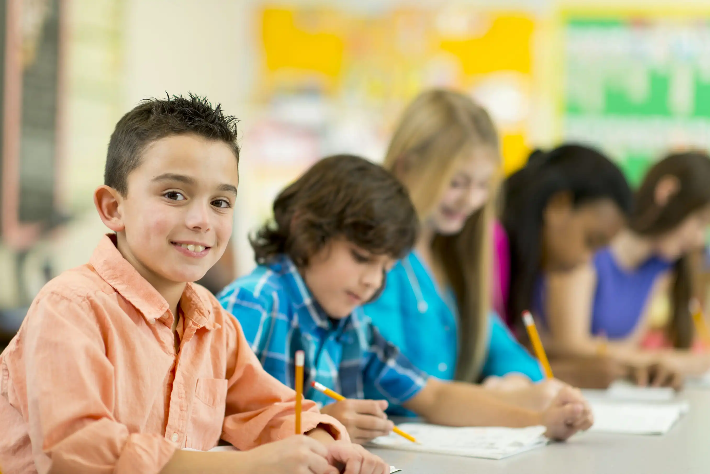A young boy in an orange shirt smiling at the camera while sitting at a classroom desk with classmates working.