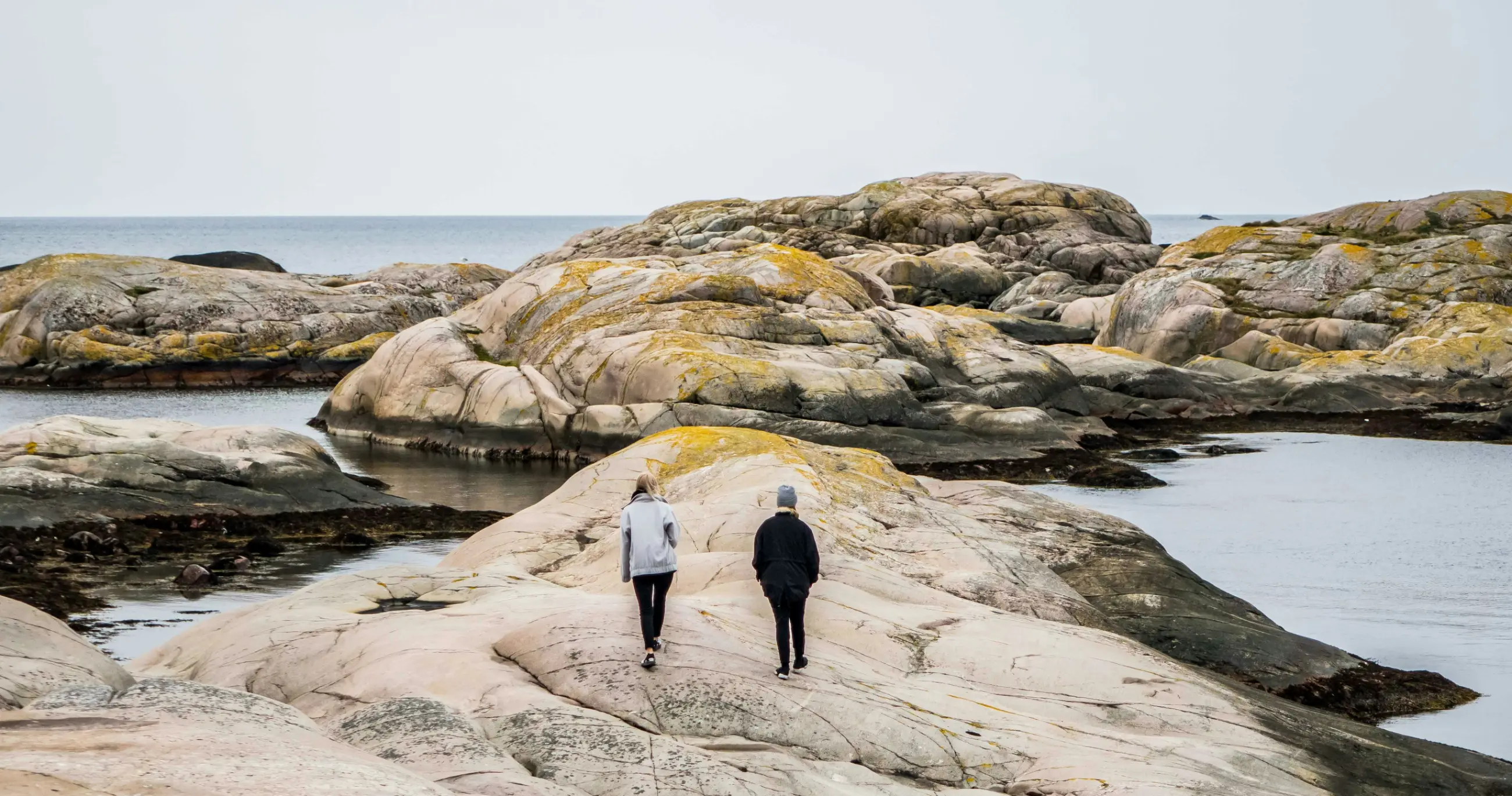 Two people walking across smooth coastal granite beside calm water on the Swedish coast