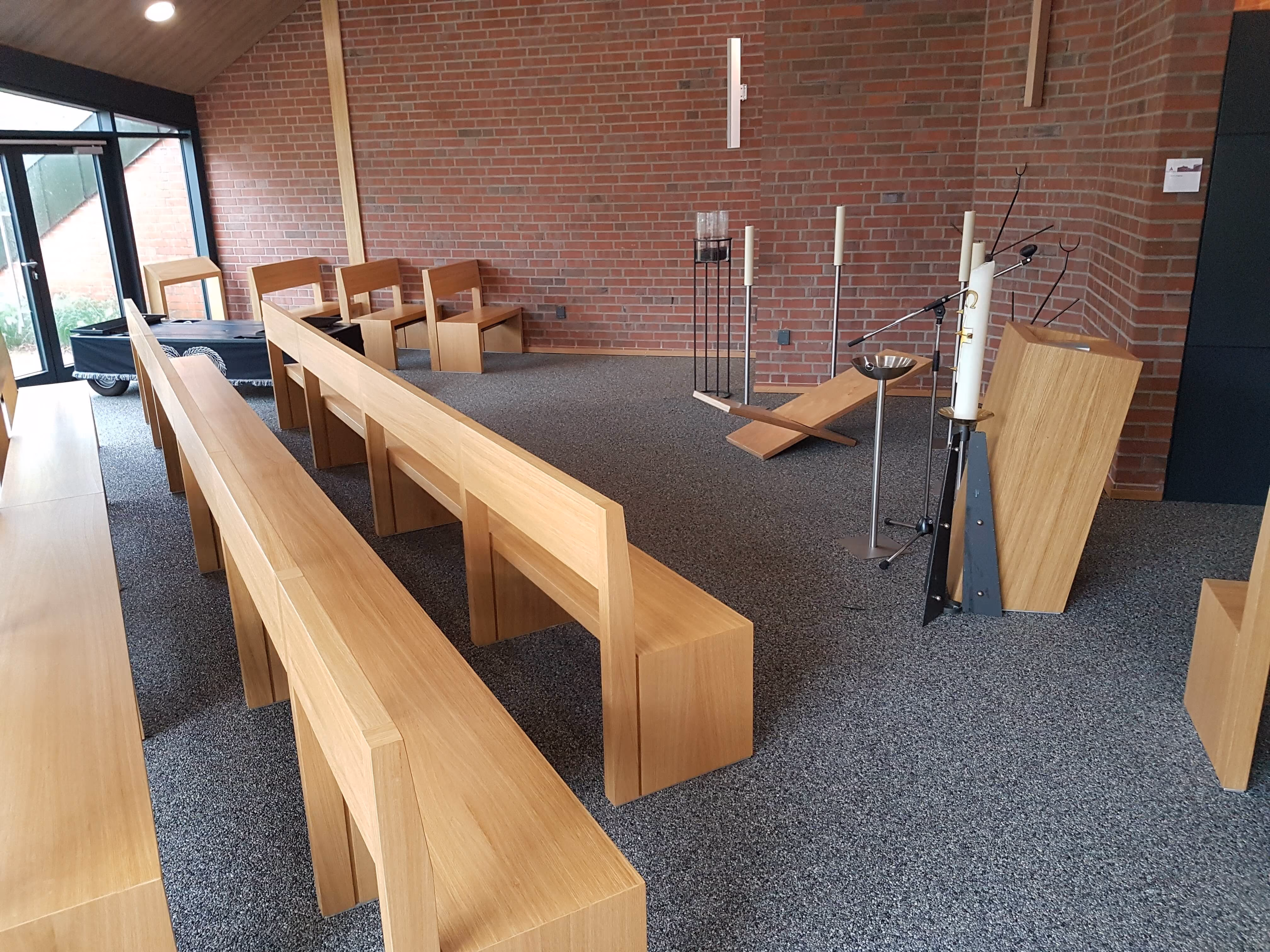 Moderner Andachtsraum mit Holzbänken, Kerzenständern, Lesepult und roter Backsteinwand.