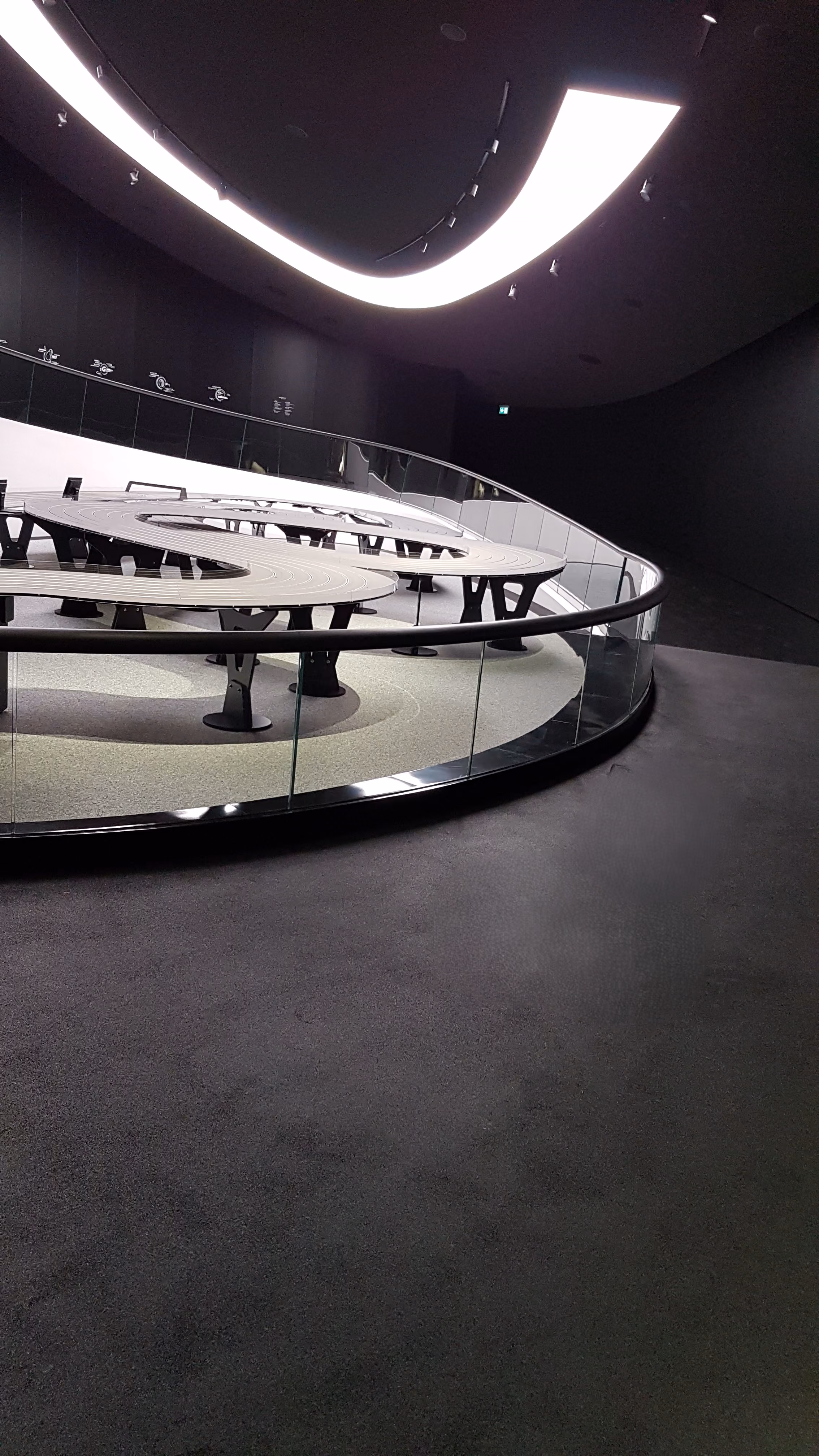 Moderne Ausstellung mit spiralförmigem Tisch und beleuchtetem Deckenelement im dunklen Raum.
