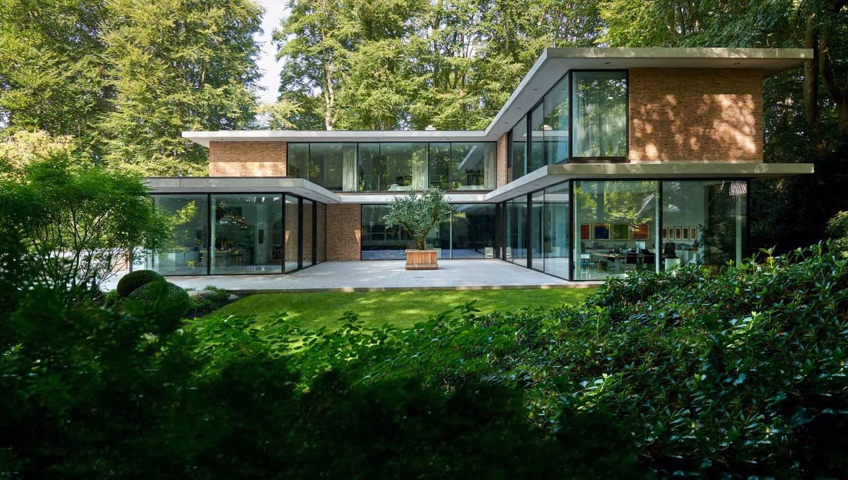Modernes Haus mit großen Glasfenstern, eingezäunter Terrasse und umgeben von Bäumen und grünen Pflanzen.
