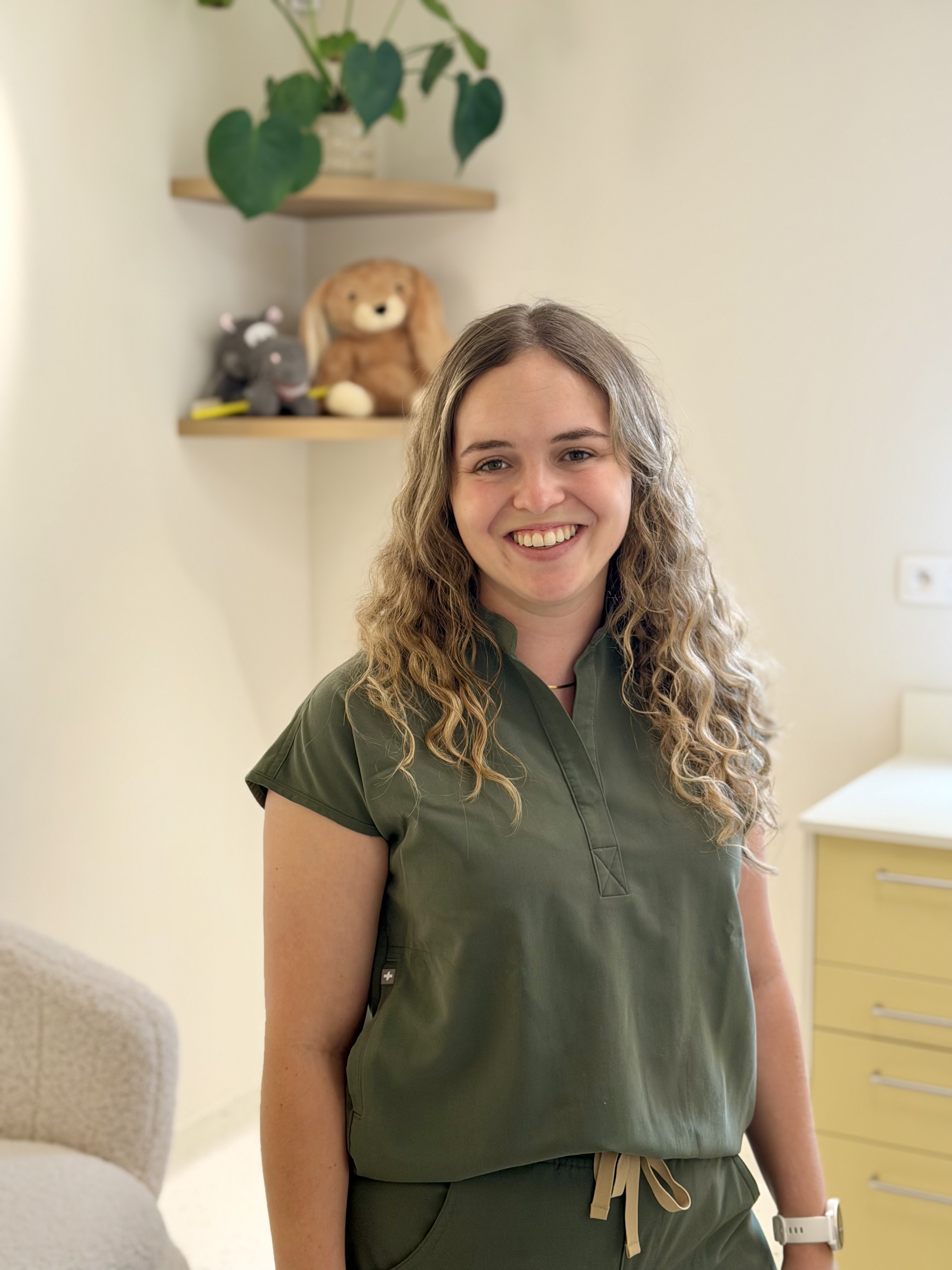 Vrouw met lang golvend blond haar draagt een groen medisch uniform en lacht naar de camera in een lichte kamer met een zachte stoel, twee houten hoekplanken met knuffels en een gele ladekast.