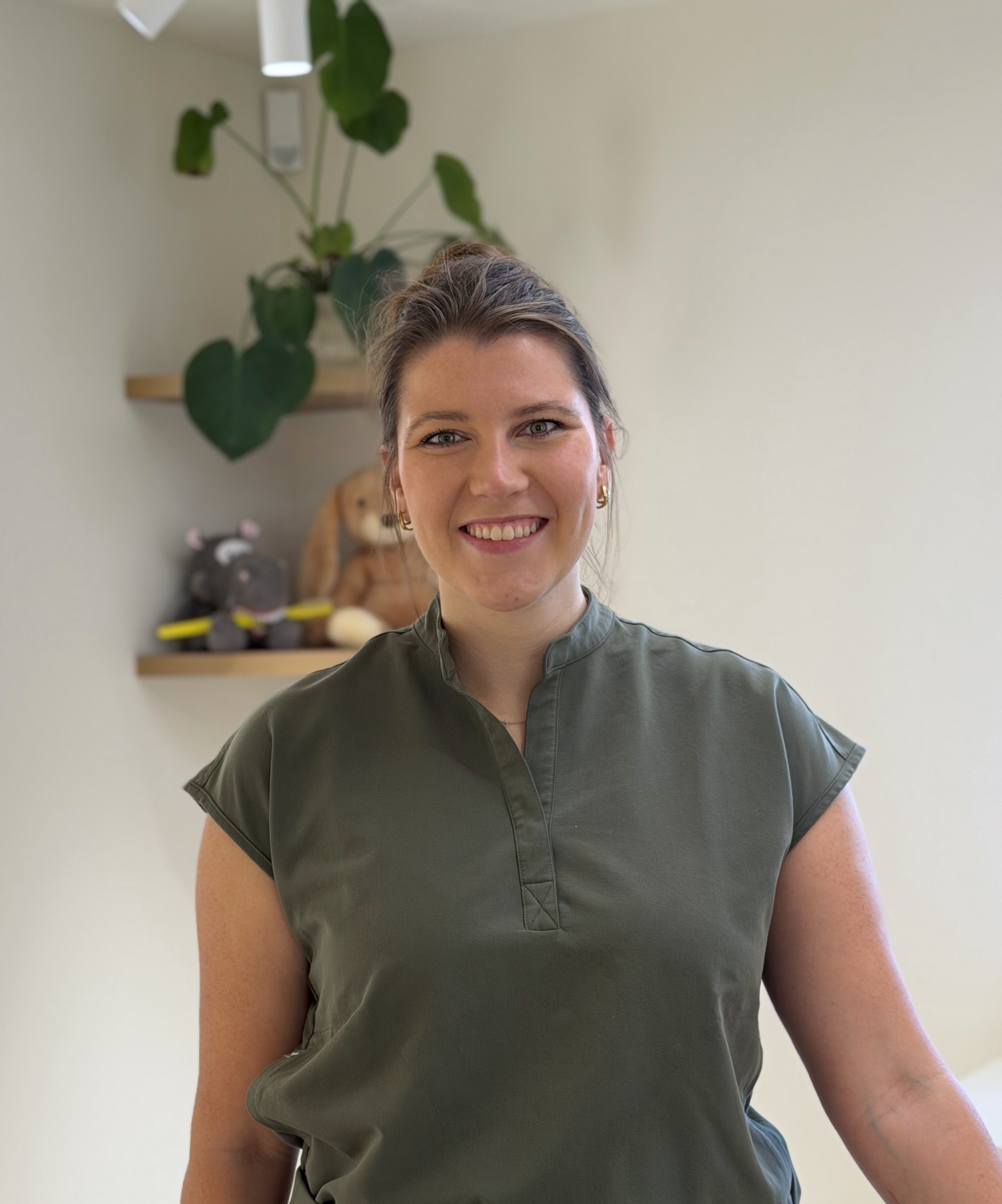 Een vrouw met een groen hemd staat glimlachend voor een witte muur met planken waarop een plant en twee knuffels staan.