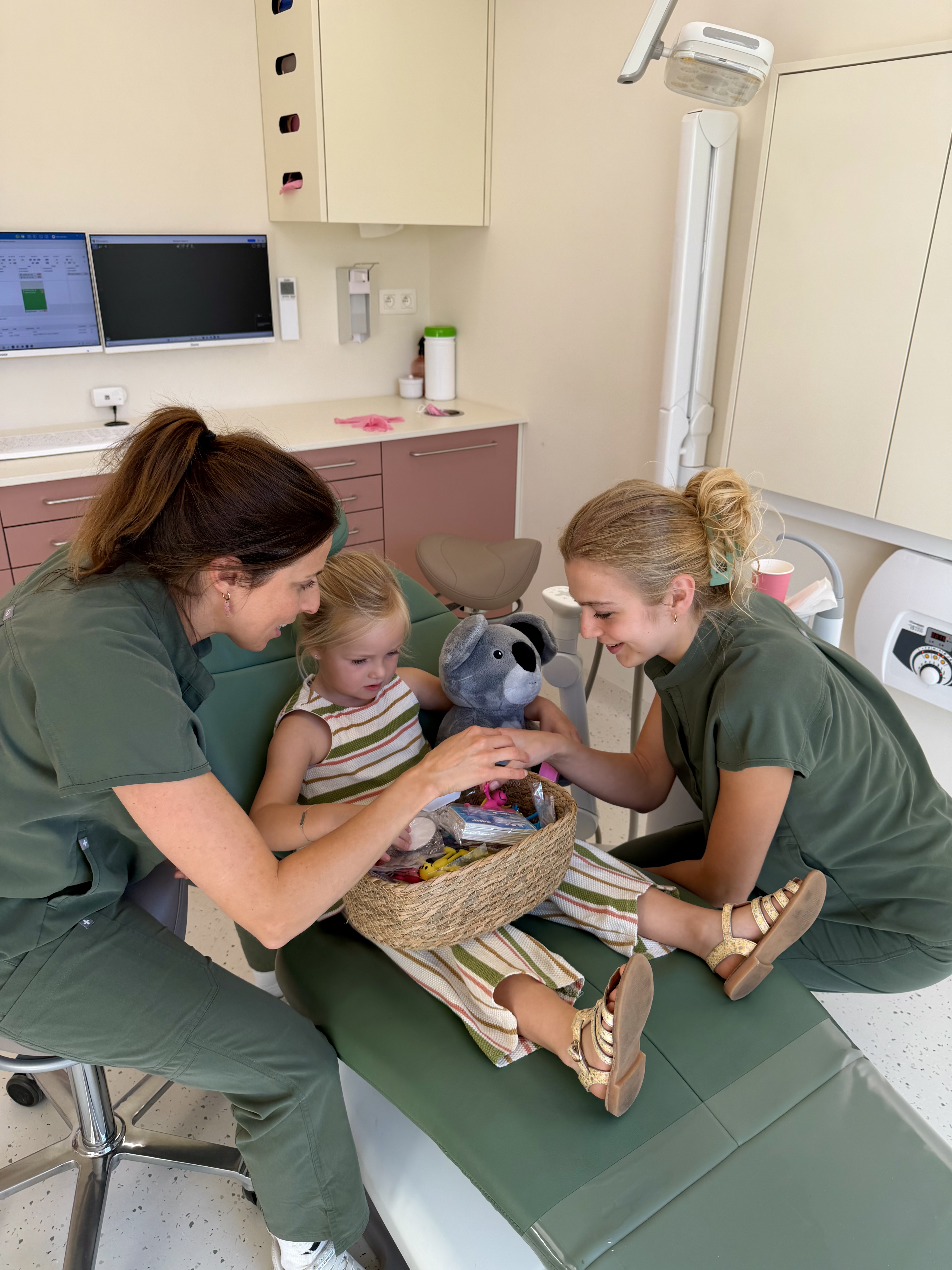 Een jong meisje zit op een groene behandelstoel in een dokterspraktijk, terwijl twee vrouwelijke zorgverleners haar aandachtig helpen en een mand met speelgoed en een knuffelkoala aanreiken.