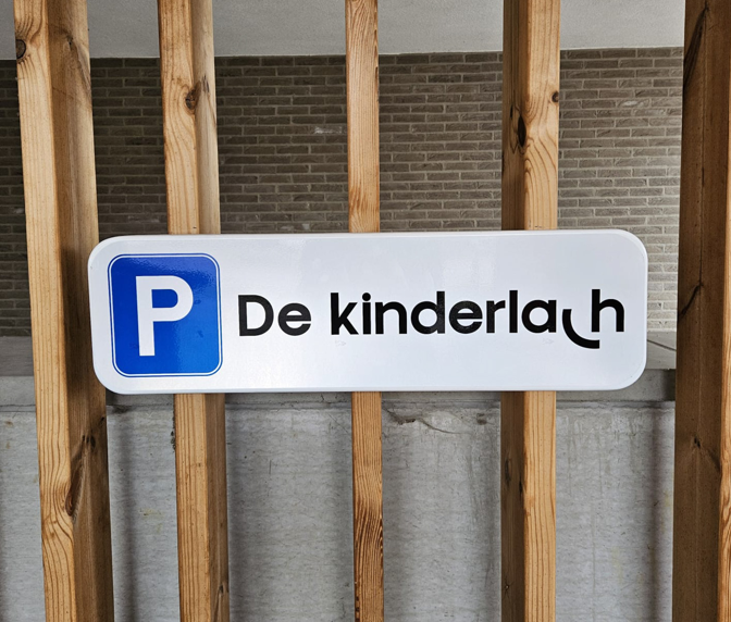 Een wit parkeerbord met een blauw pictogram P en de tekst 'De kinderlach' bevestigd aan verticale houten latten.