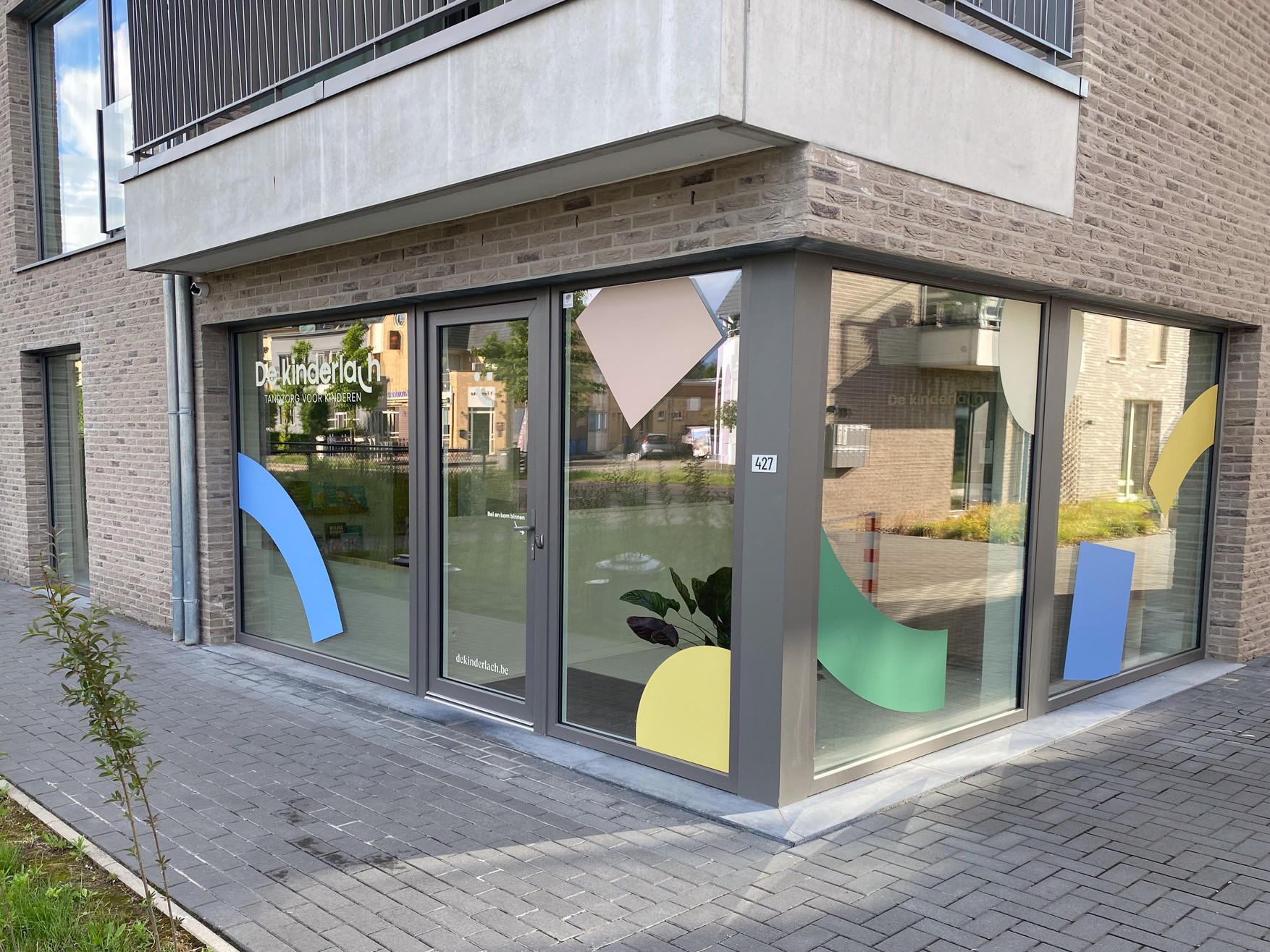 Ingang van een modern hoekgebouw met grote ramen en glazen deur met logo 'De Kinderlach', tandzorg voor kinderen, en kleurrijke geometrische vormen op de ramen.