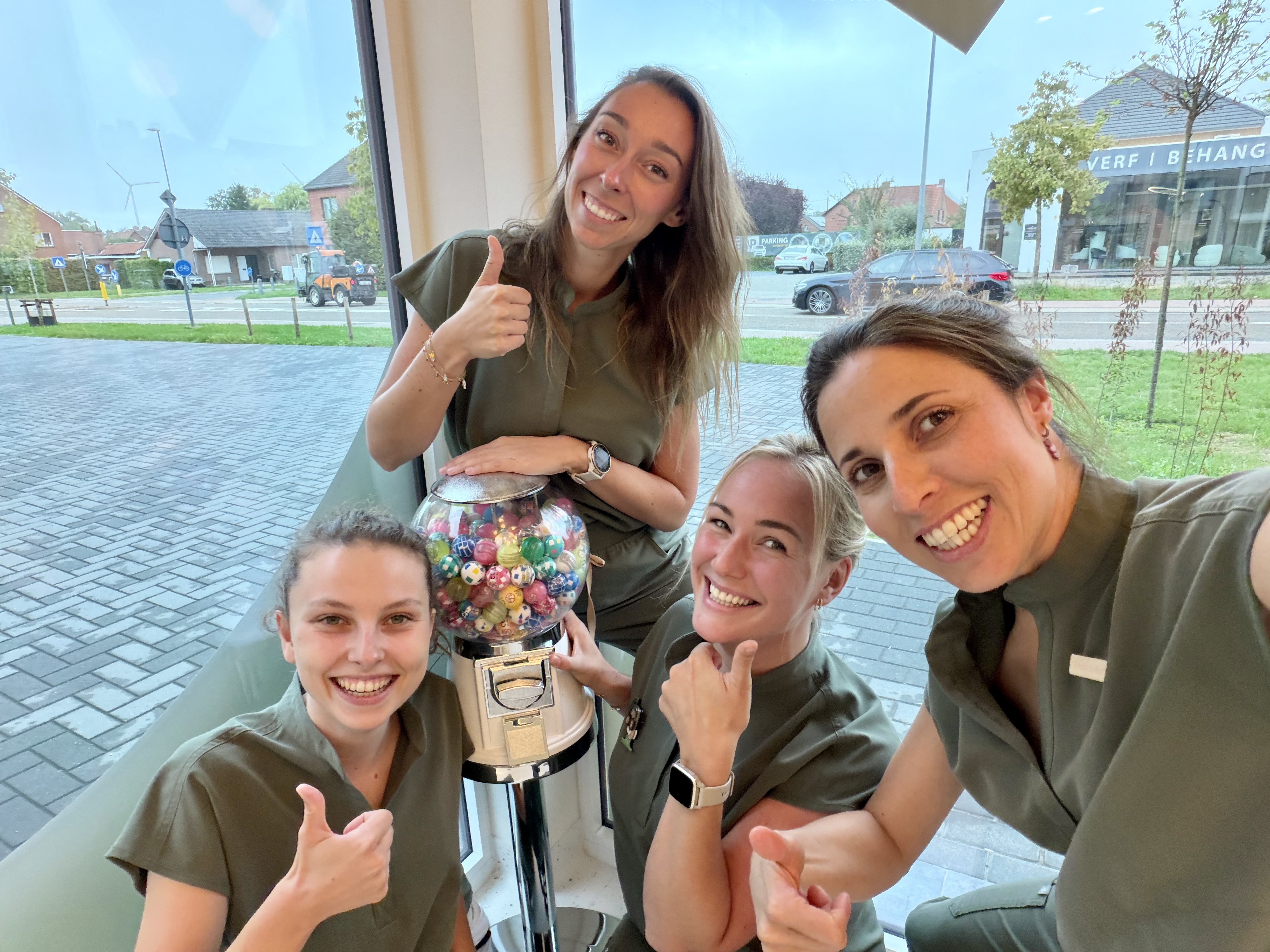 Vier glimlachende vrouwen in groene uniformen poseren samen bij een raam met uitzicht op een straat; ze tonen allemaal een duim omhoog en staan rond een snoepautomaat gevuld met kleurrijke balletjes.