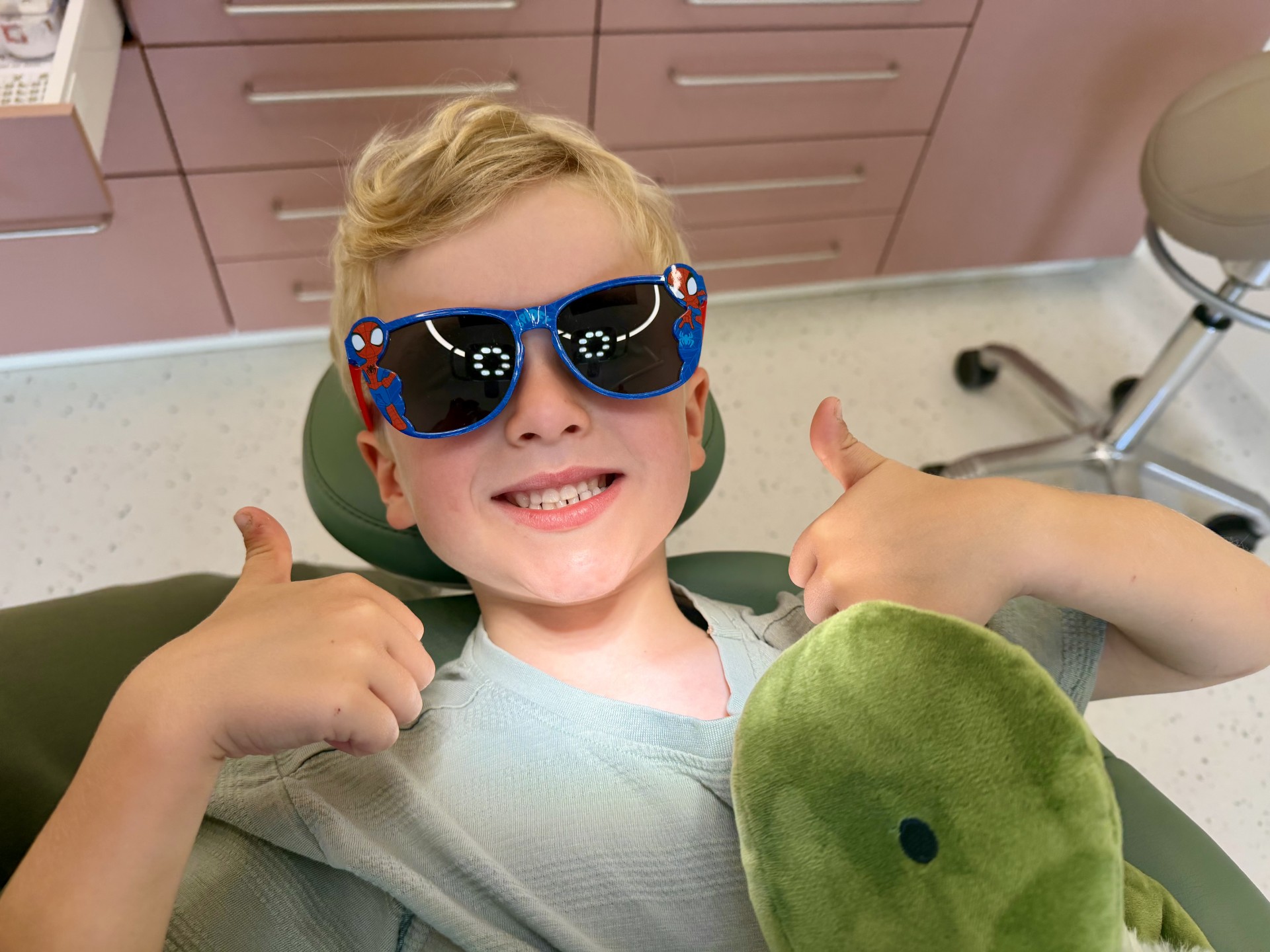 Jongen met blonde krullen zit in een stoel, draagt Spider-Man-zonnebril en geeft met beide handen een duim omhoog, terwijl hij een groene knuffel vasthoudt.