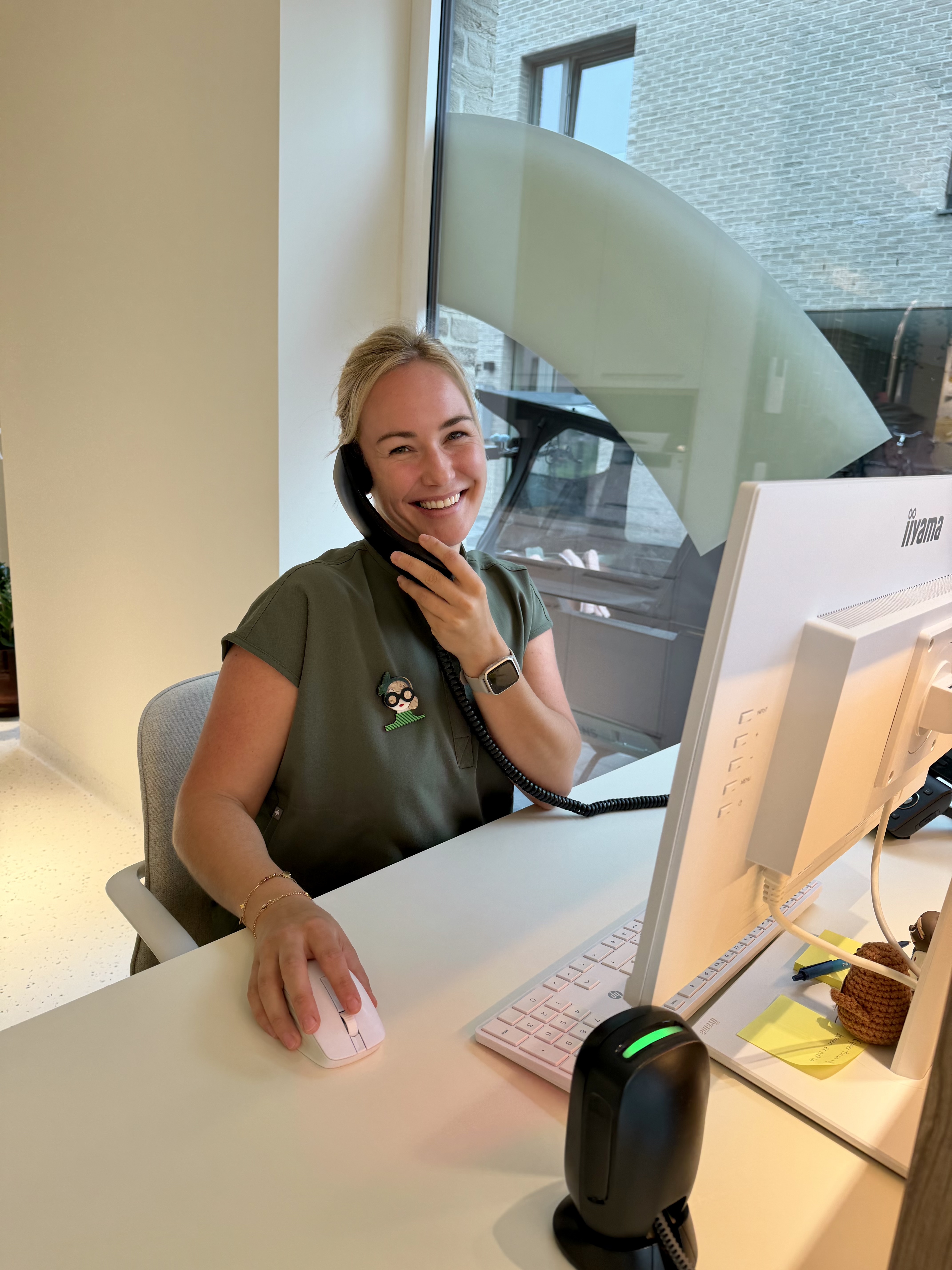 Vrouw met blond haar lacht en spreekt aan de telefoon terwijl ze aan een bureau zit, met haar hand op een computermuis, achter een computerscherm bij een raam.