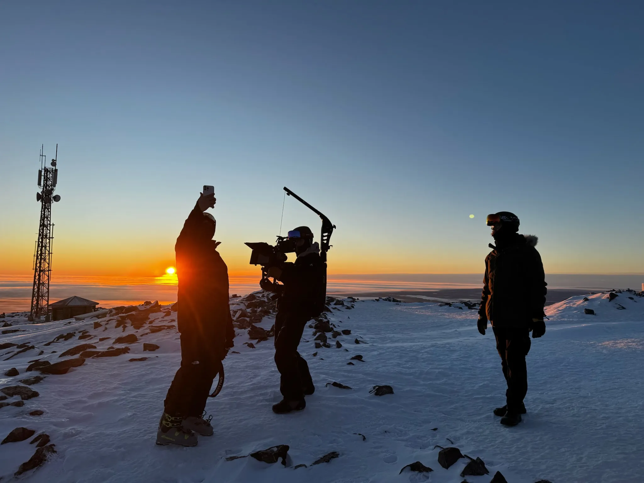 Kolme kuvausryhmän jäsentä lumisella tunturilla auringonlaskussa.