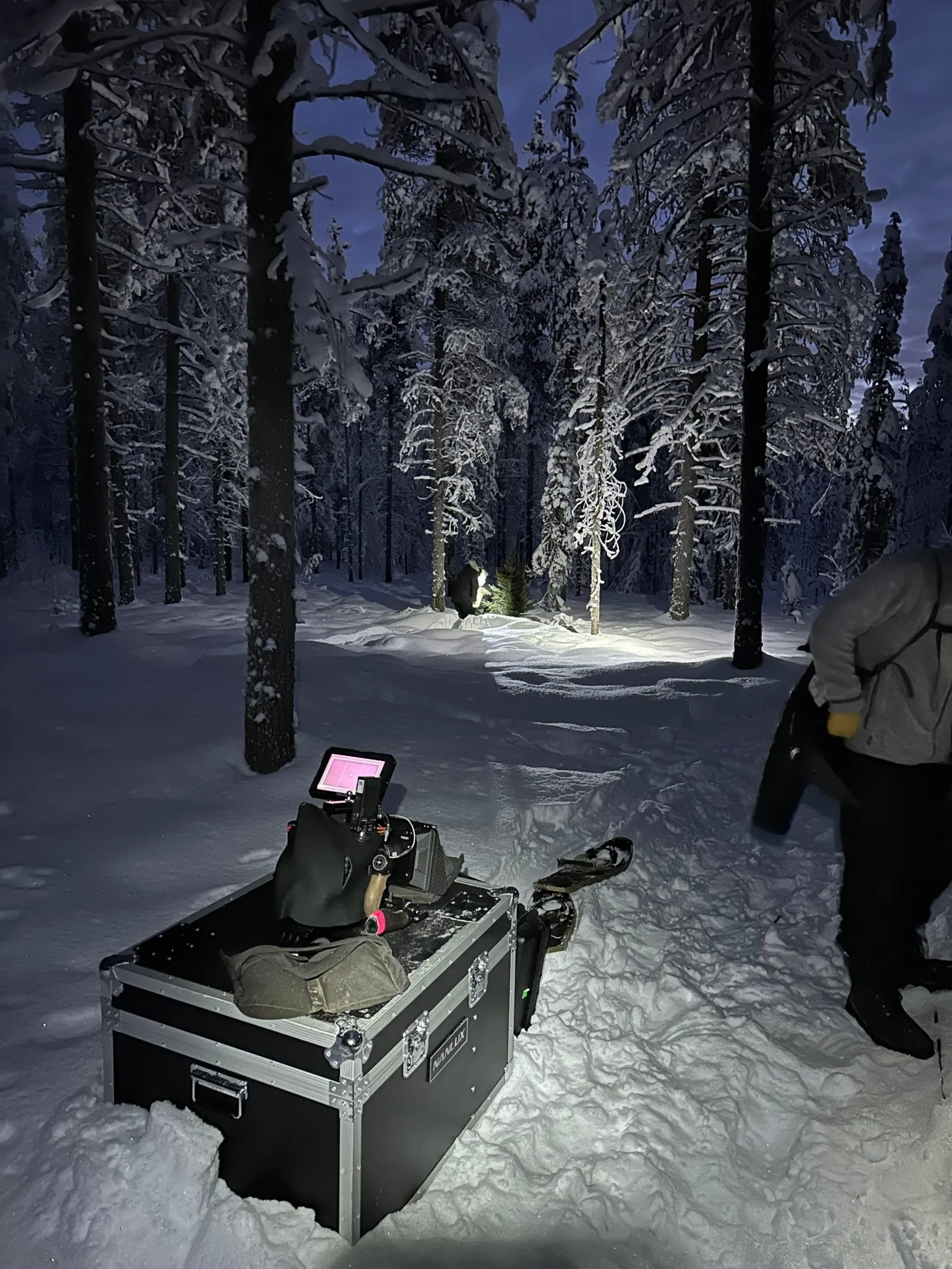 A camera and crew in an illuminated snow-covered forest at dusk.