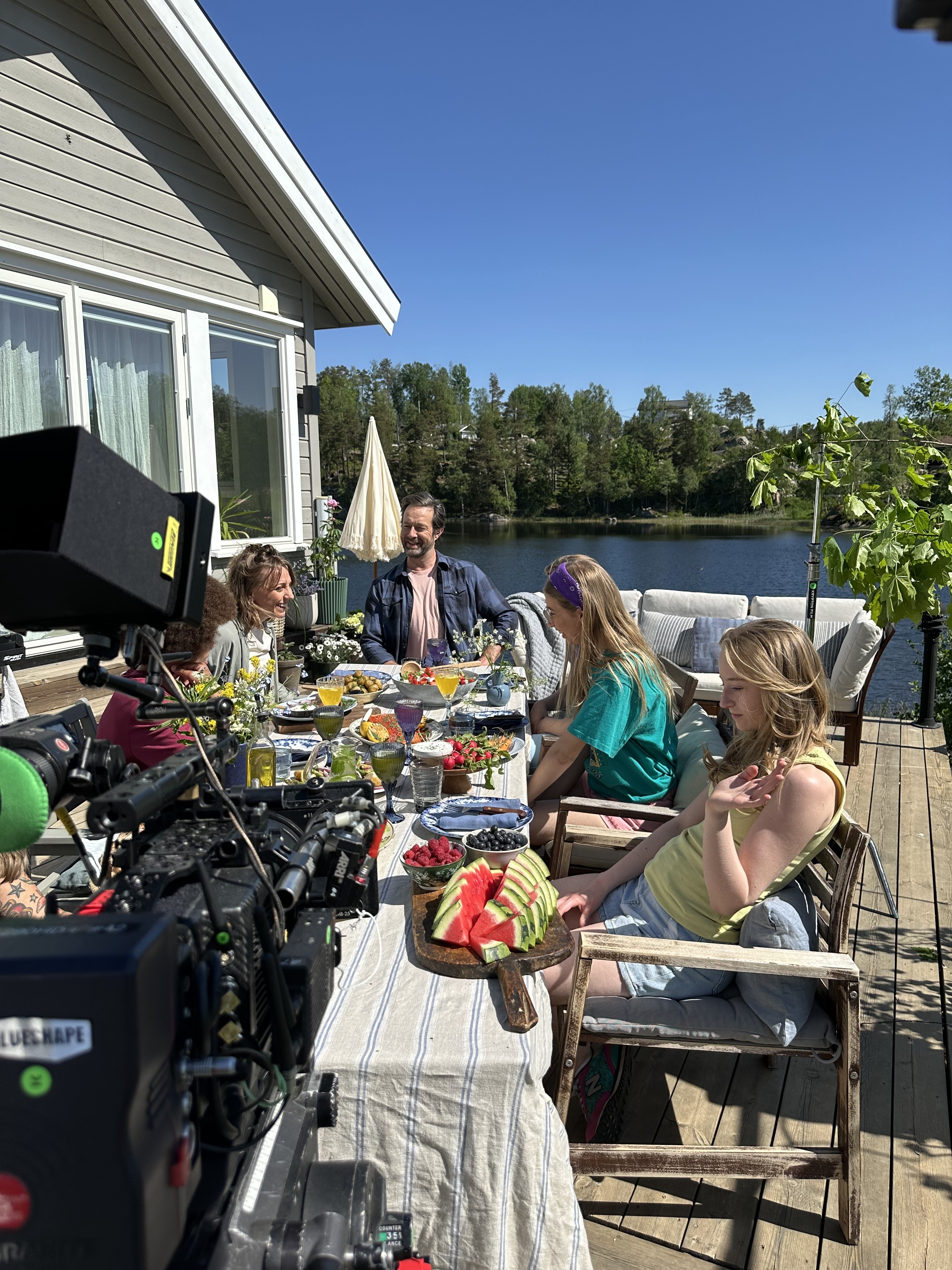Kamera ja esiintyjät kesäisen ruokapöydän äärellä järvenrantaterassilla S-marketin kuvauksissa.