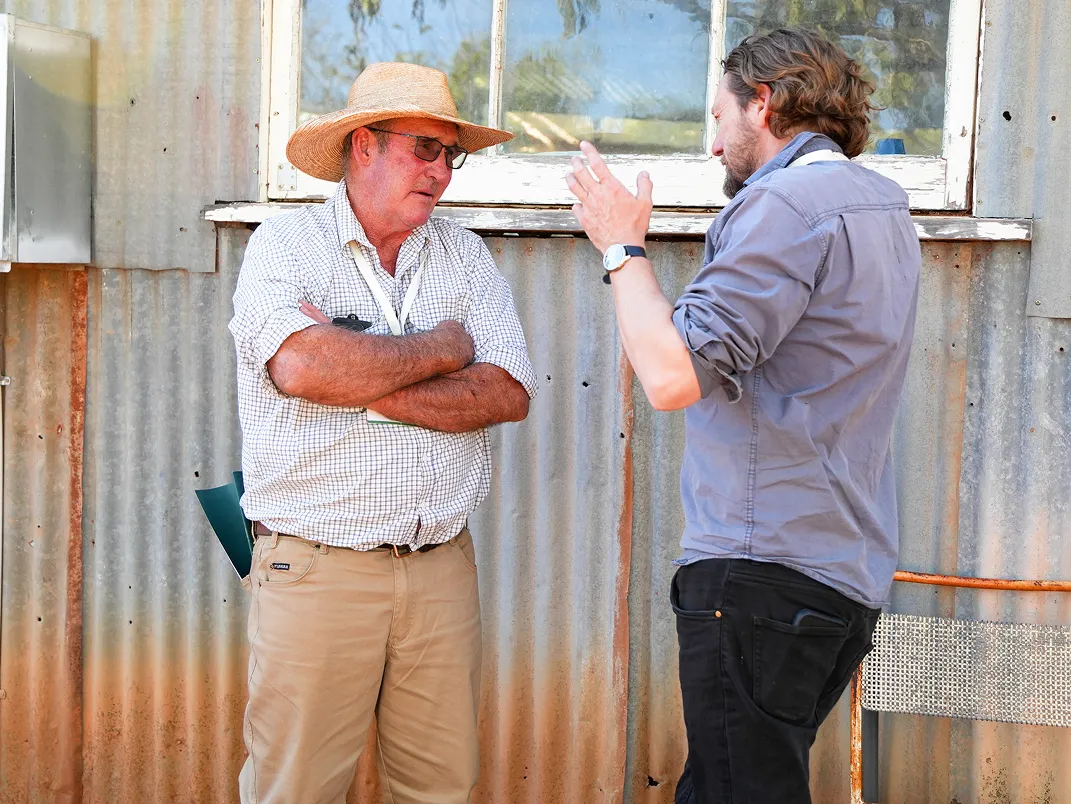 Two men having a conversation outdoors in front of a corrugated metal wall with a window; one man wears a straw hat, glasses, a checkered shirt, and khaki pants with arms crossed, while the other man with rolled-up sleeves gestures with his hands.