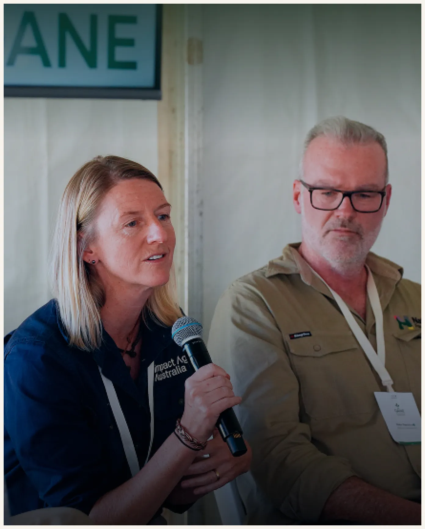 A woman with blonde hair wearing a dark blue shirt with an 'Impact Net Australia' logo speaks into a microphone, while a man with glasses and gray hair in a beige shirt listens beside her.
