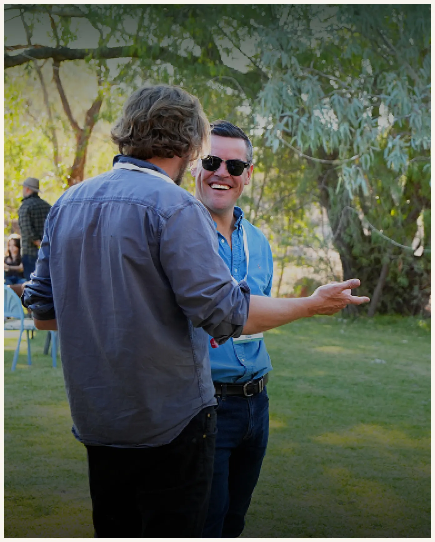 Two men standing outdoors on grass having a conversation; one facing the camera wearing sunglasses and smiling, the other with back to camera gesturing with his hand.