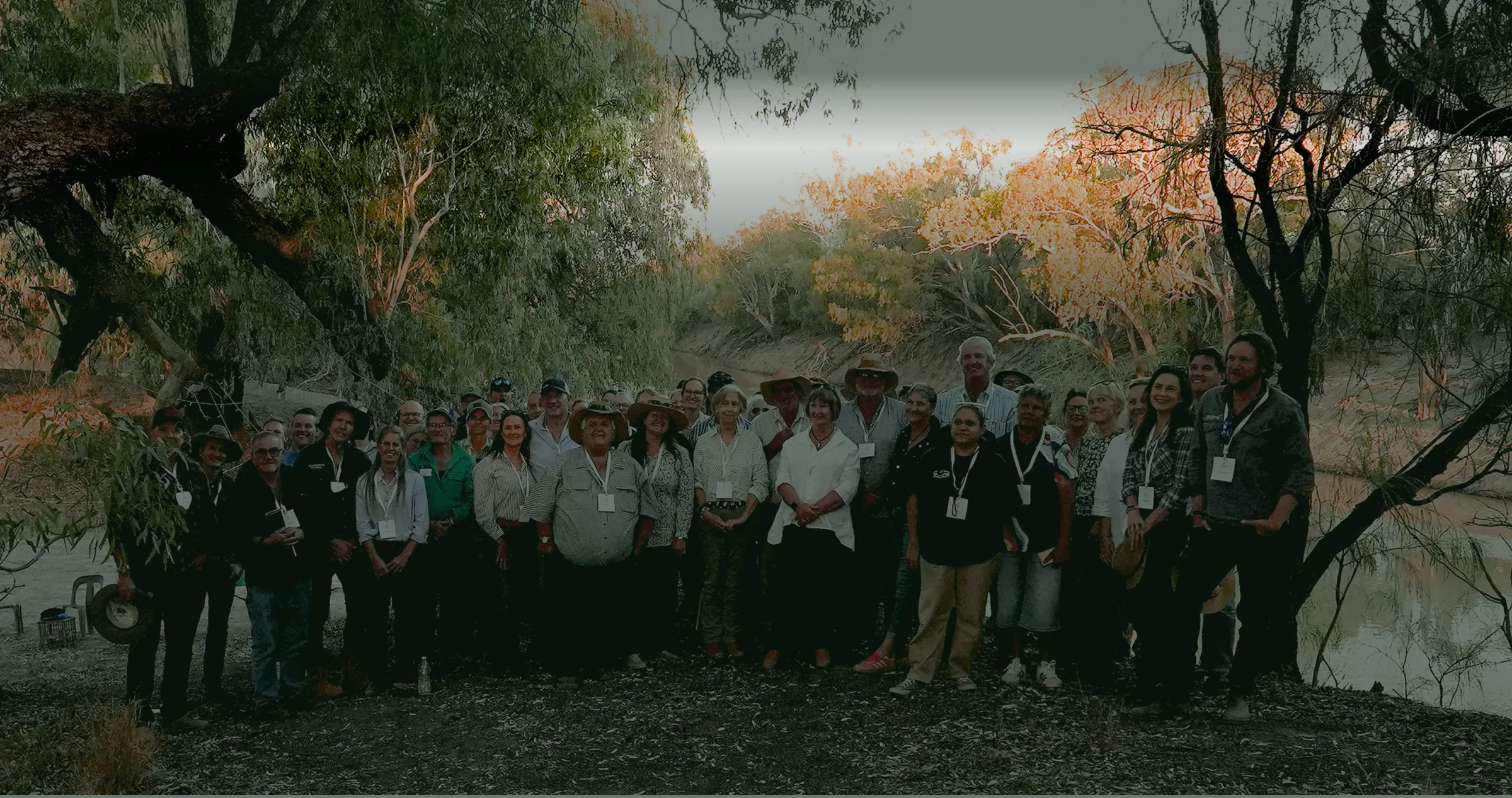 Large group of adults standing together outdoors in a wooded area near a riverbank during sunset, with trees and foliage surrounding them.