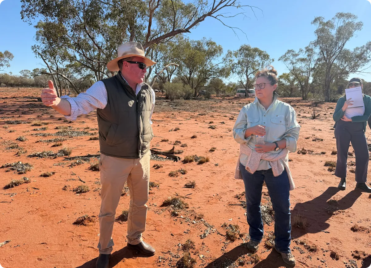 Man wearing a wide-brimmed hat, sunglasses, and a vest gesturing with his left hand while talking to a woman holding a water bottle in a dry, reddish dirt outdoor area with scattered vegetation and trees under a clear blue sky; another woman stands in the background holding papers.