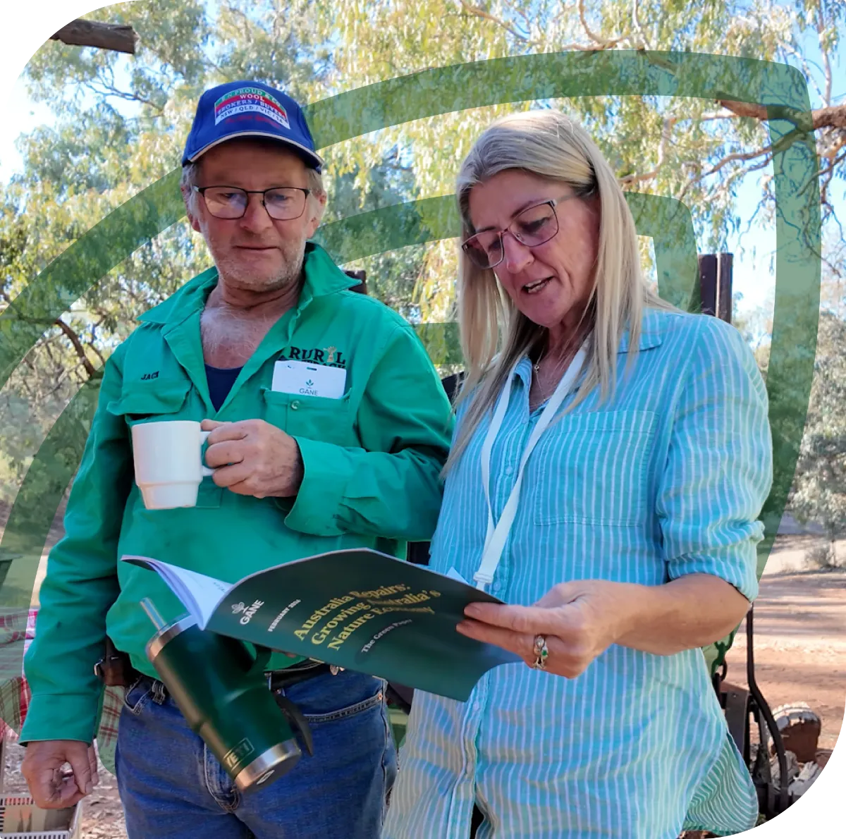 A man wearing a green shirt and blue cap holds a white mug while a woman in a light blue striped shirt and glasses reads a booklet titled 'Australia Begins: Growing Australia's Nature Recovery'. They are outdoors with trees in the background.