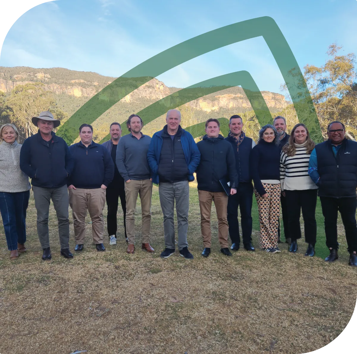A group of twelve adults standing outdoors on grass with mountains and trees in the background on a clear day. They are dressed in casual warm clothing including jackets, sweaters, and scarves, and they are facing the camera.