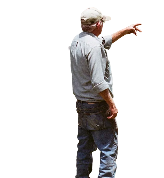 Older man wearing a beige cap, light blue shirt with rolled-up sleeves, and jeans, standing and pointing to the right with his right arm extended.