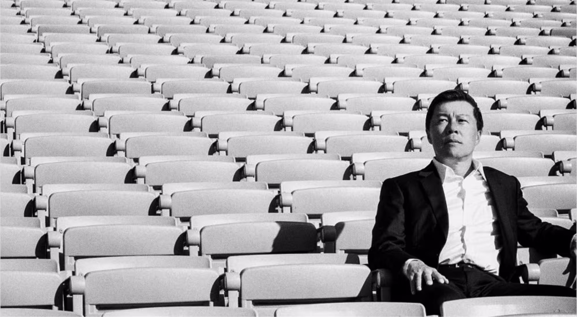 Man in a dark suit sitting alone in an empty stadium with rows of empty seats behind him.