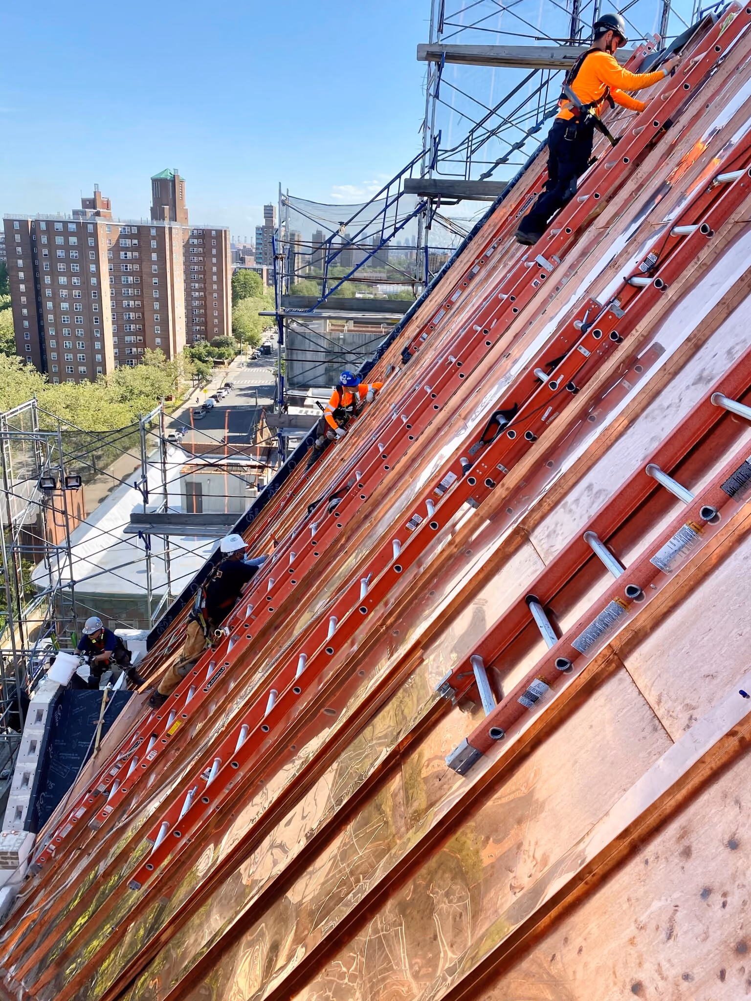 Construction workers in safety gear working on a steep, framed roof with ladders in an urban area.