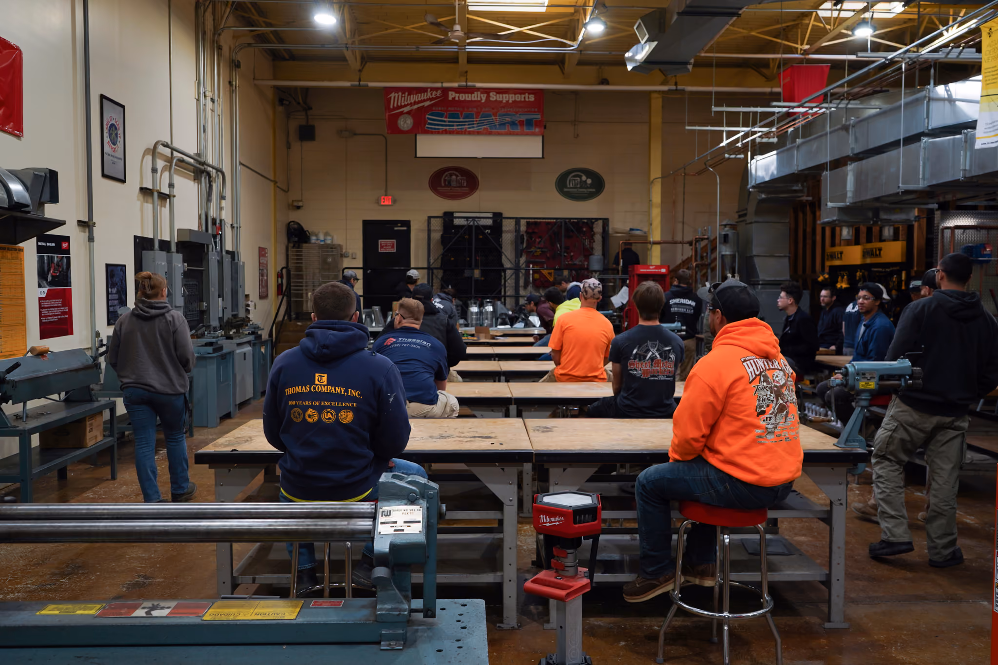 People seated and standing in a workshop or classroom with tools and machines around.
