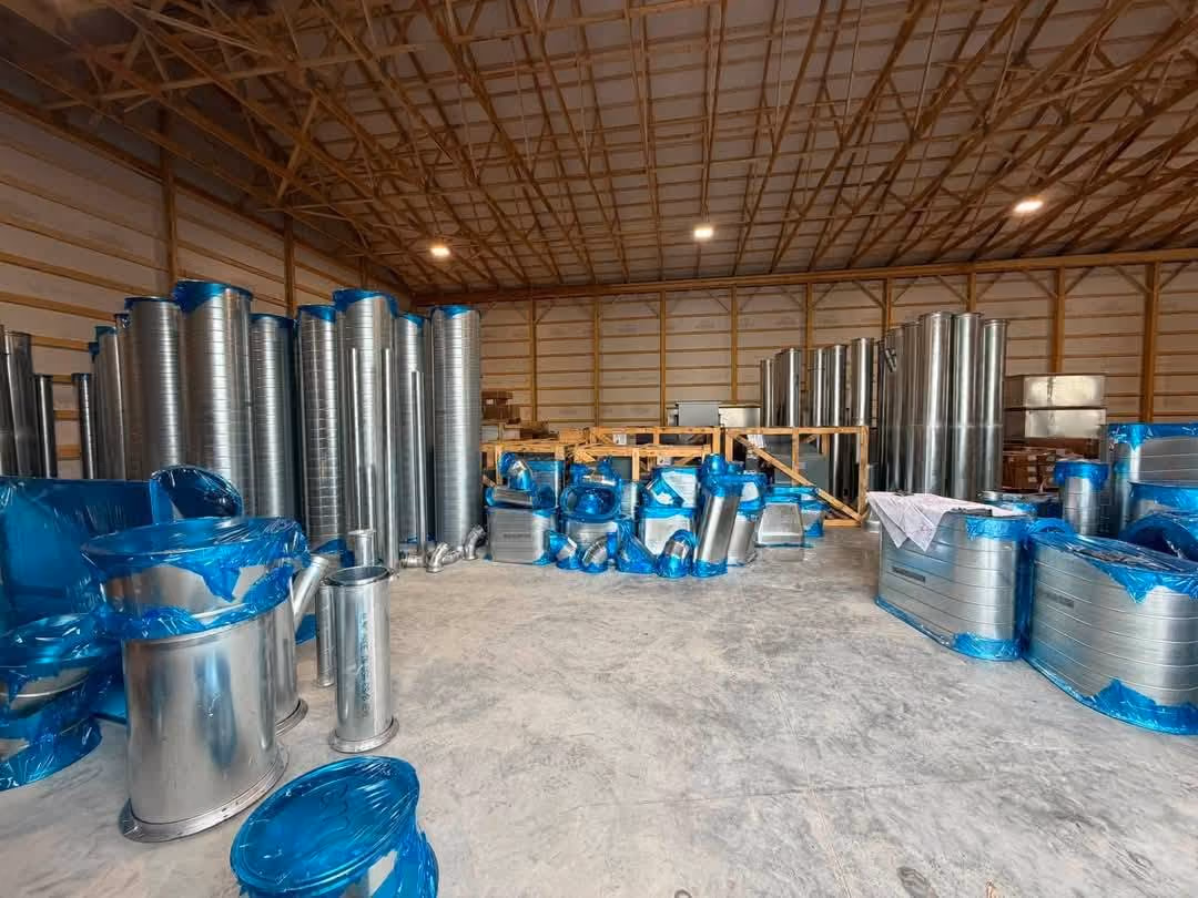 Metal HVAC ductwork parts wrapped in blue plastic stored inside a large warehouse with wooden beams.
