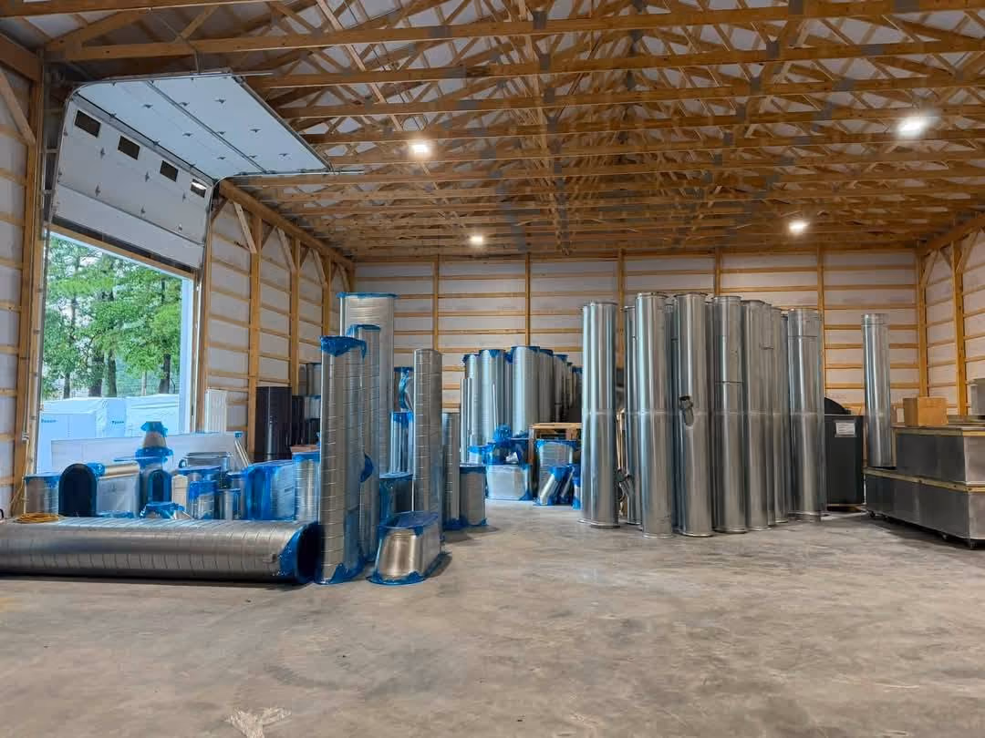 Metal HVAC ducts and components stacked inside a large wooden warehouse with an open garage door.