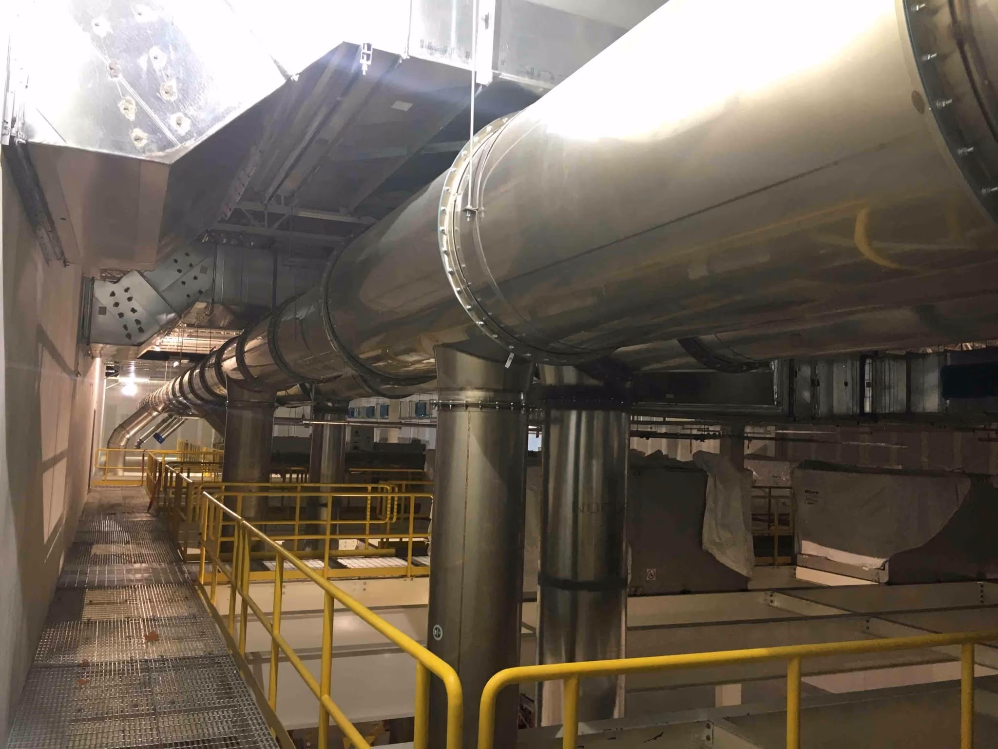 Industrial ductwork system with large metal pipes and yellow railings inside a facility.