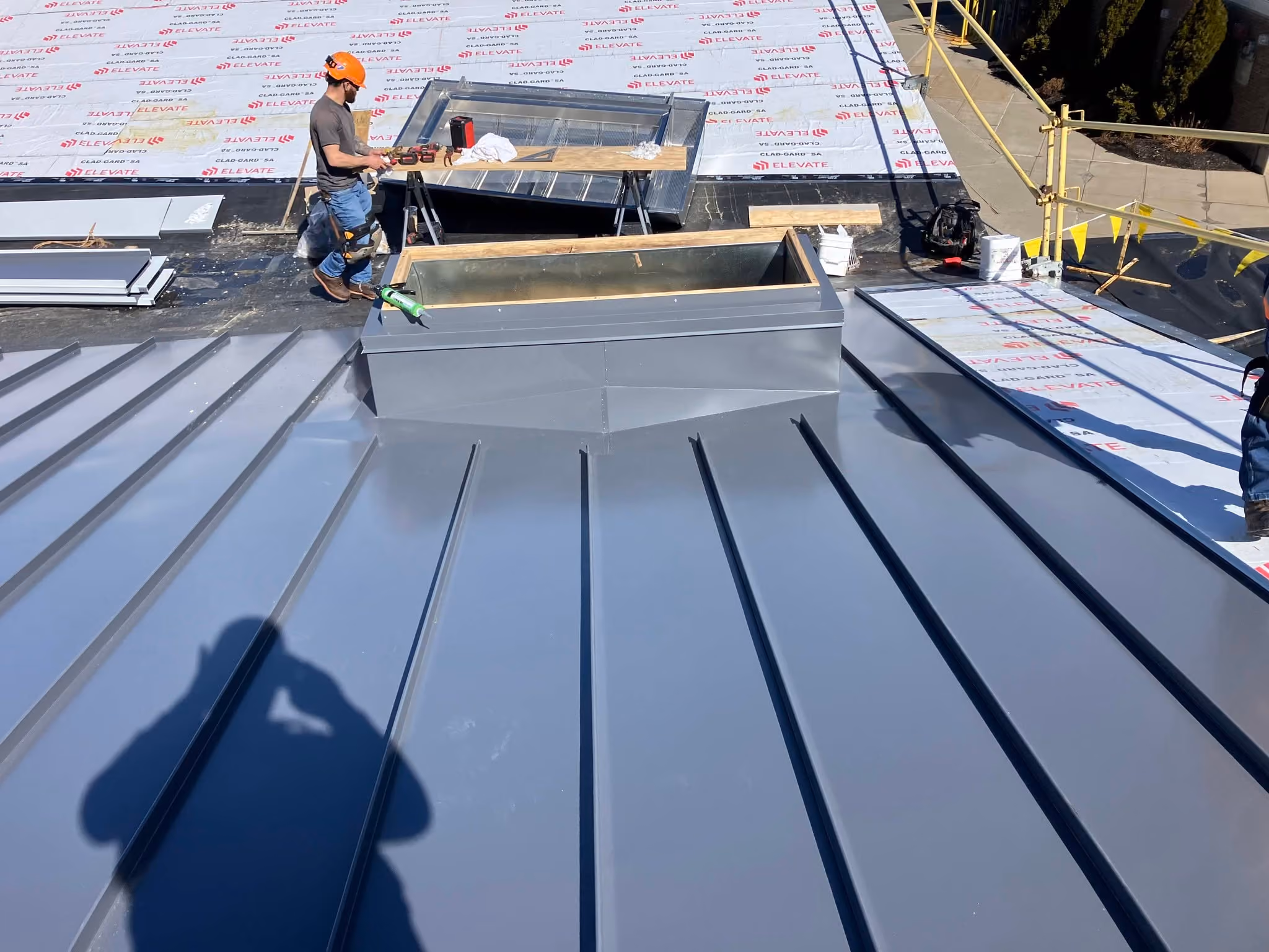 Worker on a roof under construction near a gray standing seam metal panel and skylight opening.