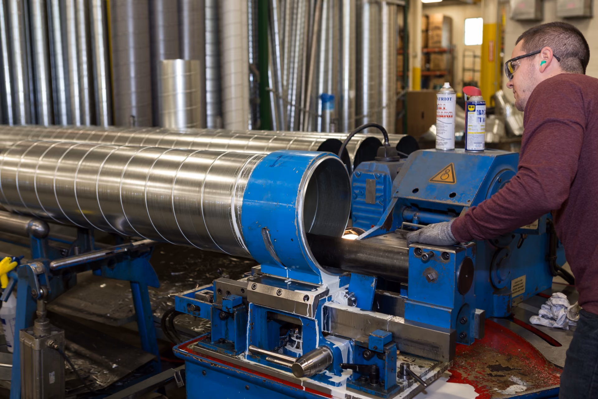 Worker wearing safety glasses and earplugs operates blue metalworking machine with large metal pipe.